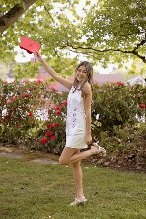 Smiling female graduate in white dress and 2026 sash joyfully raises red cap while posing in garden.