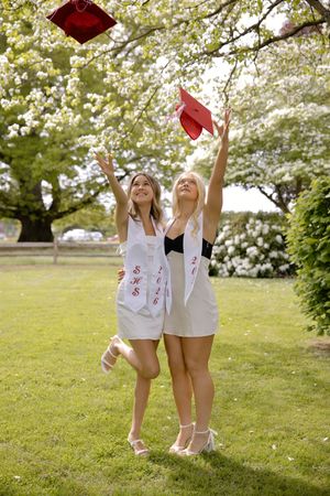 Two female graduates in white dresses toss red graduation caps into the air beneath a blossoming tree.