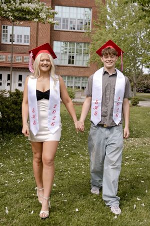 Two high school graduates in red caps and white 2026 stoles hold hands outdoors near a brick school building.