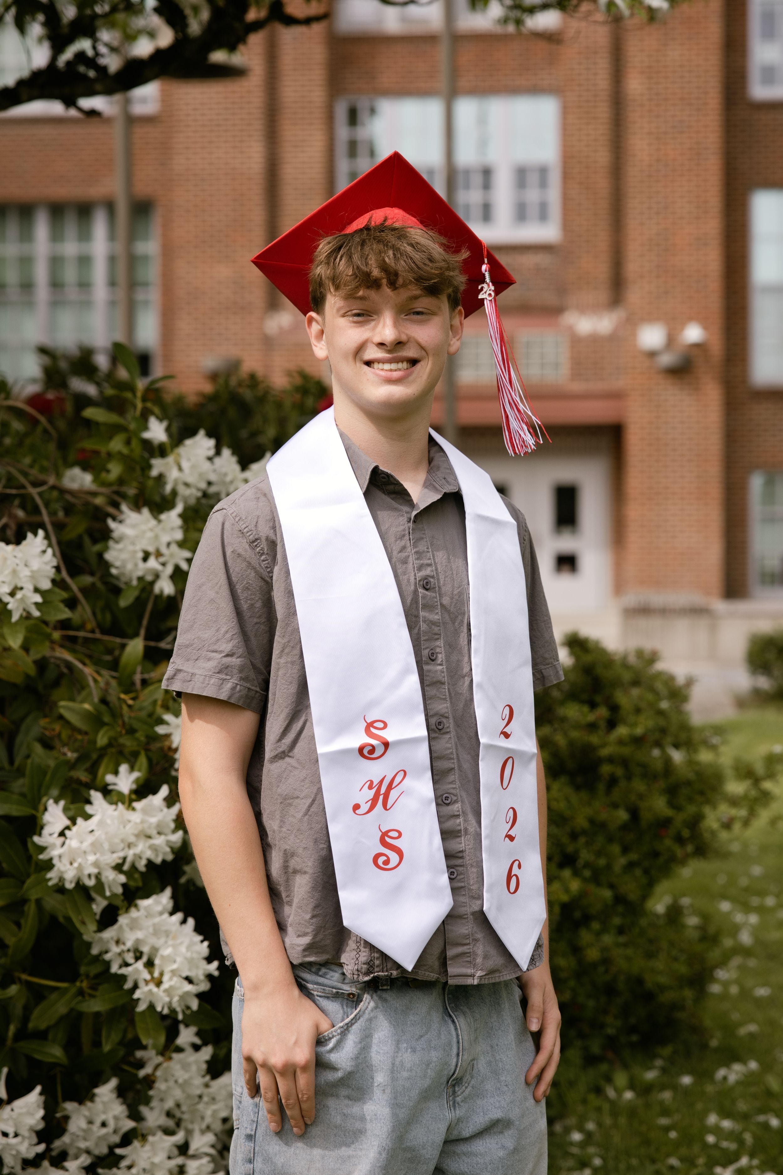 Smiling high school graduate wearing red cap and white SHS 2026 stole poses outdoors near flowering shrubs.