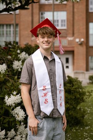 Smiling high school graduate wearing red cap and white SHS 2026 stole poses outdoors near flowering shrubs.