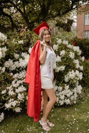 A smiling graduate poses in a red cap and gown over a white dress in front of white flowering bushes.