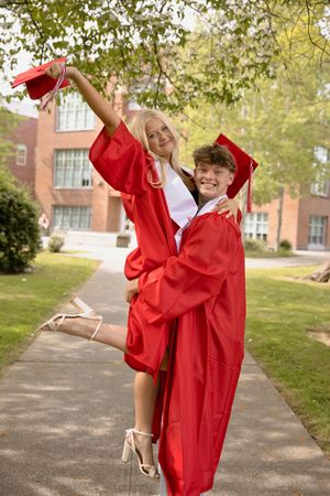 Two graduates in red caps and gowns celebrate outdoors, one lifting the other joyfully on a school walkway.