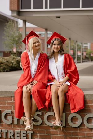 Two female graduates in red caps and gowns sit together on a brick wall outside their high school.