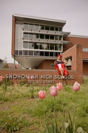 Graduate in red cap and gown sits on Snohomish High School sign with tulips in foreground.