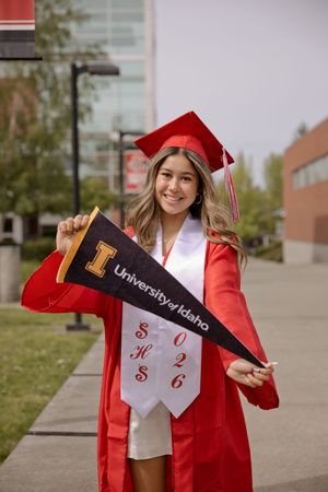 Smiling female graduate in red cap and gown holding University of Idaho pennant on campus.