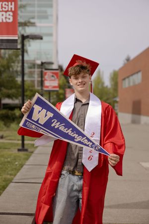Smiling graduate in red cap and gown holding a University of Washington pennant and sash outdoors.