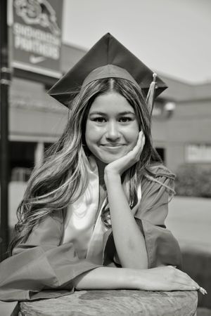 Black and white portrait of a female graduate in cap and gown posing thoughtfully outside her school.