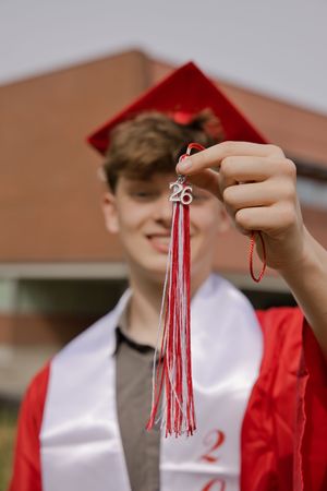 A male graduate in red cap and gown holds up a 2026 graduation tassel outside a brick building.
