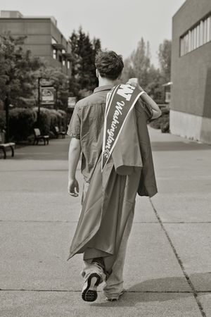 Male graduate walking away in gown with Washington sash on urban campus in black and white.