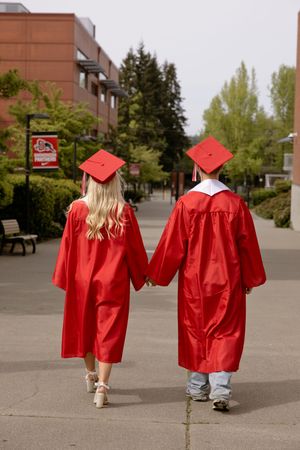 Two graduates in red caps and gowns walking hand in hand down a campus pathway.