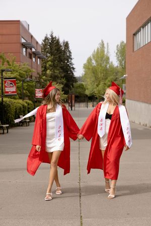 Two female graduates in red caps and gowns hold hands while walking on a campus pathway.