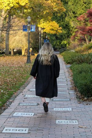Blonde woman in black graduation gown walks down brick pathway lined with year markers on a colorful autumn campus.