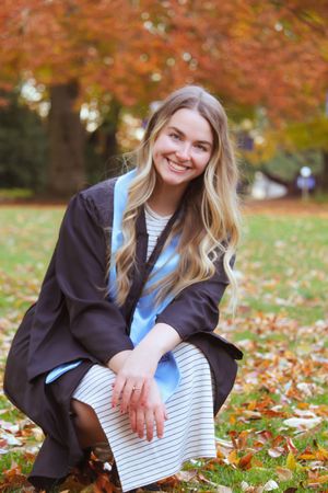Smiling blonde woman in graduation gown and blue sash crouching among autumn leaves in a park.