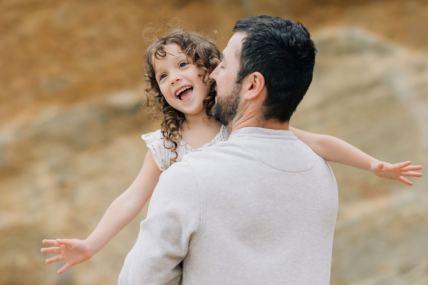 A joyful little girl with curly hair spreads her arms wide while being held by her bearded father outdoors.