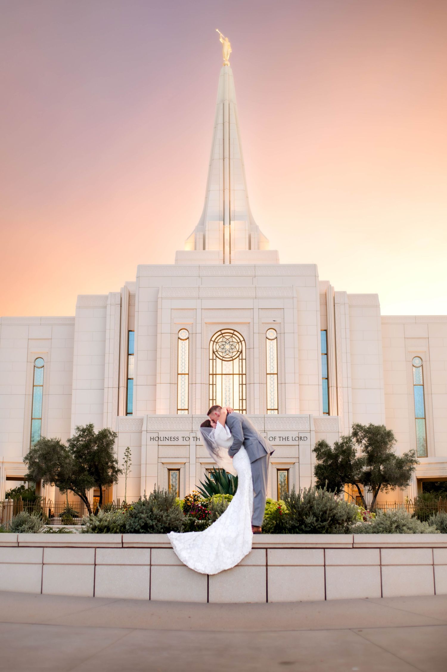 Groom dips bride in a romantic kiss in front of an illuminated LDS temple at sunset.