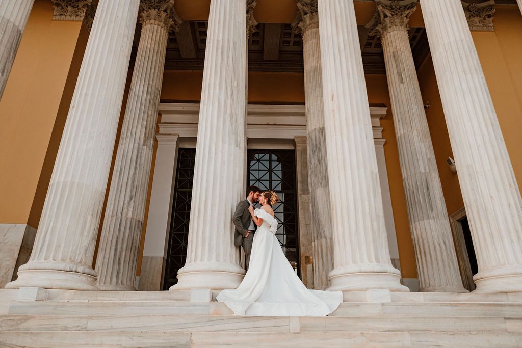 Bride and groom embracing on grand neoclassical building steps with tall white columns during outdoor wedding photoshoot.