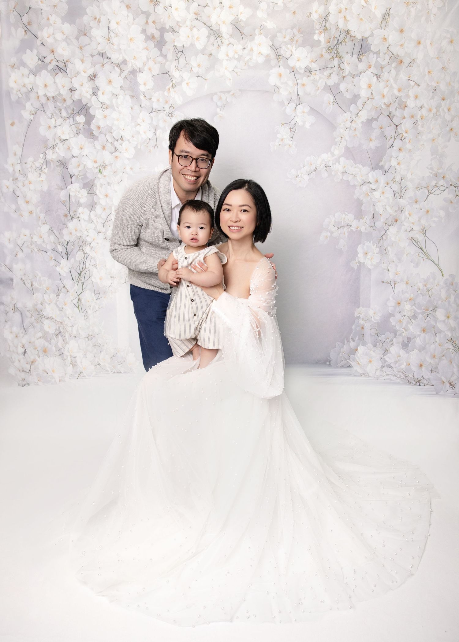 Family portrait of parents and baby in white floral studio, mother in elegant white gown, father in gray sweater.