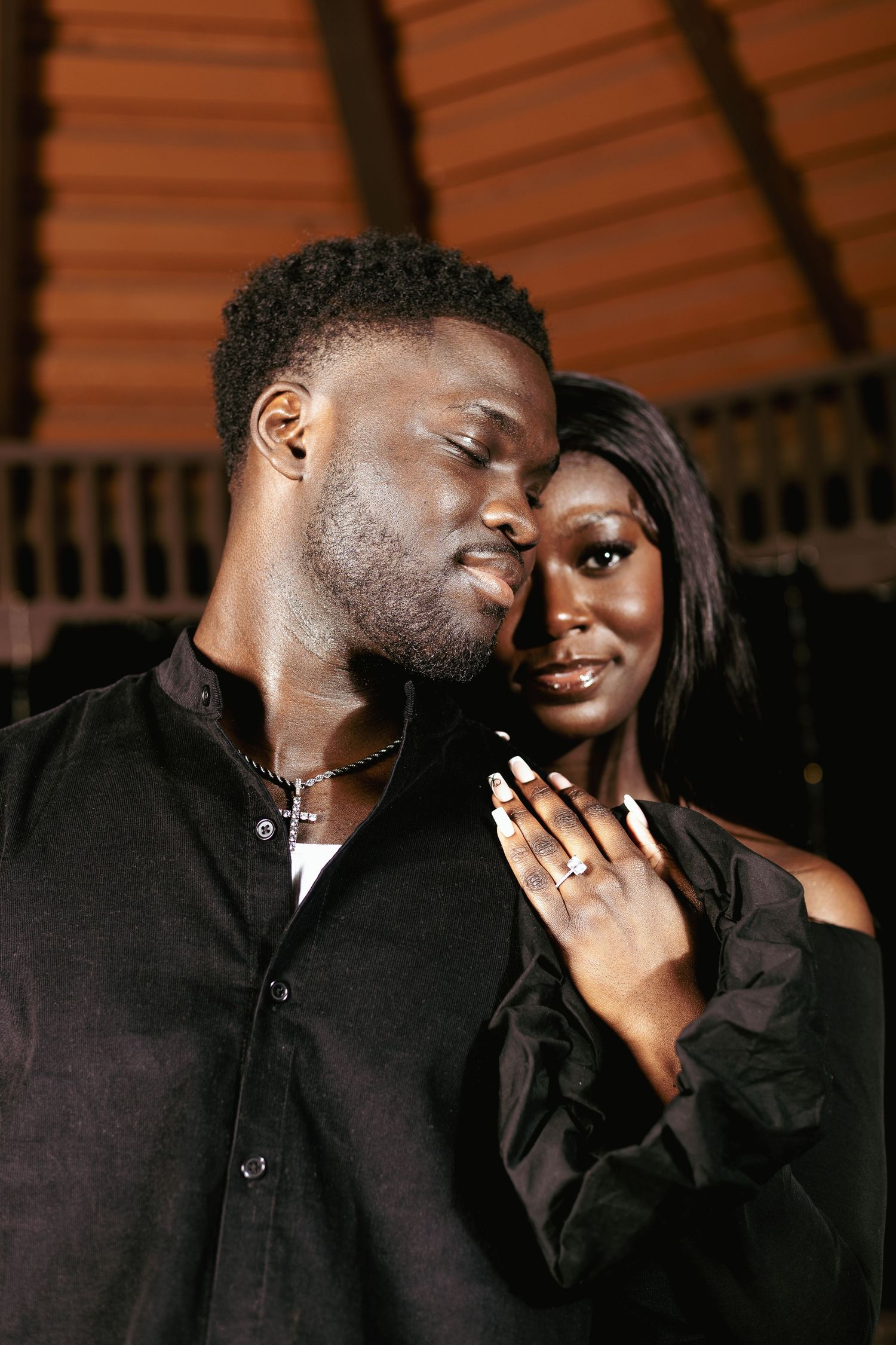 An elegant Black couple in matching black outfits pose closely together in a warmly lit intimate portrait.