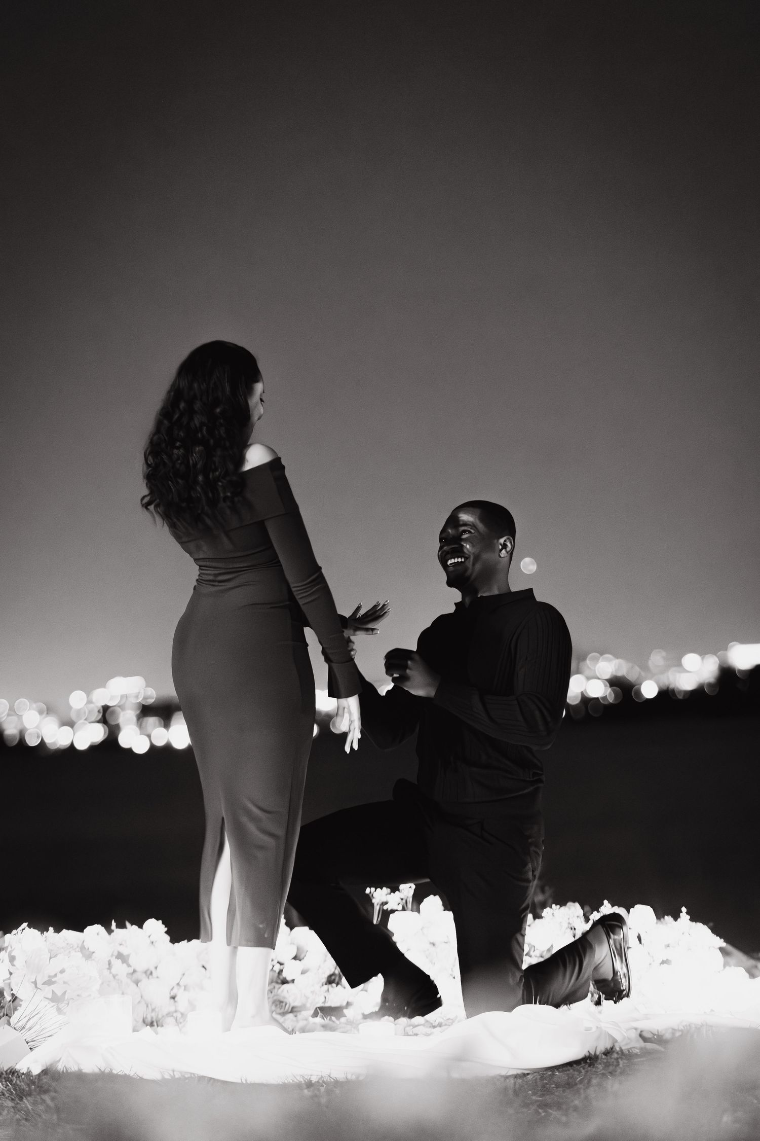 A man proposes on one knee to a woman against a city lights backdrop in a romantic black and white night photo.