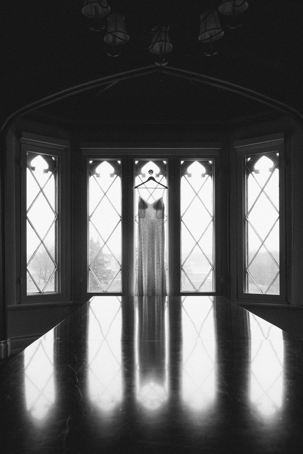 Black and white photo of a wedding dress hanging in a gothic bay window, reflecting on polished floor.