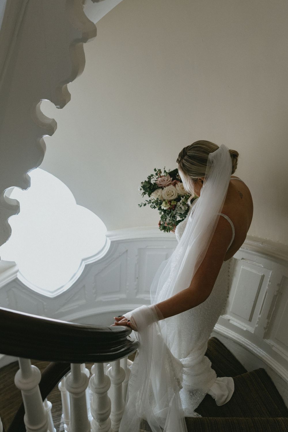 Bride in white gown and veil descends ornate staircase holding a pastel floral bouquet in an elegant venue.