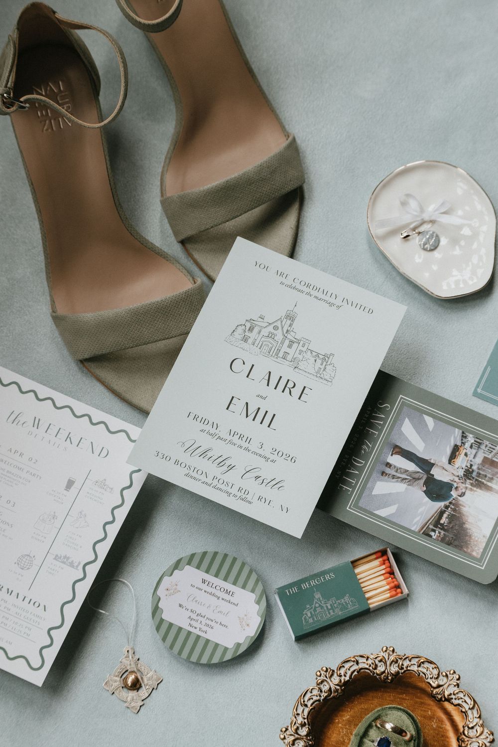 Elegant wedding flat lay with sage green heels, floral invitation for Claire and Emil, matches, and jewelry on a mint background.