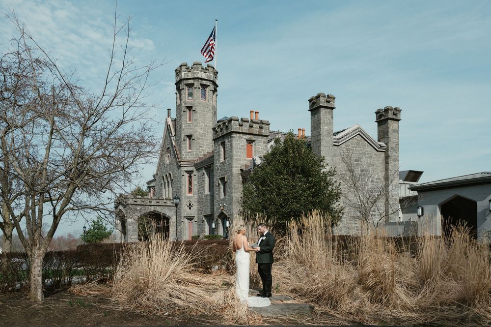 Couple poses before a stone castle venue with American flag, surrounded by winter landscape at a romantic wedding.