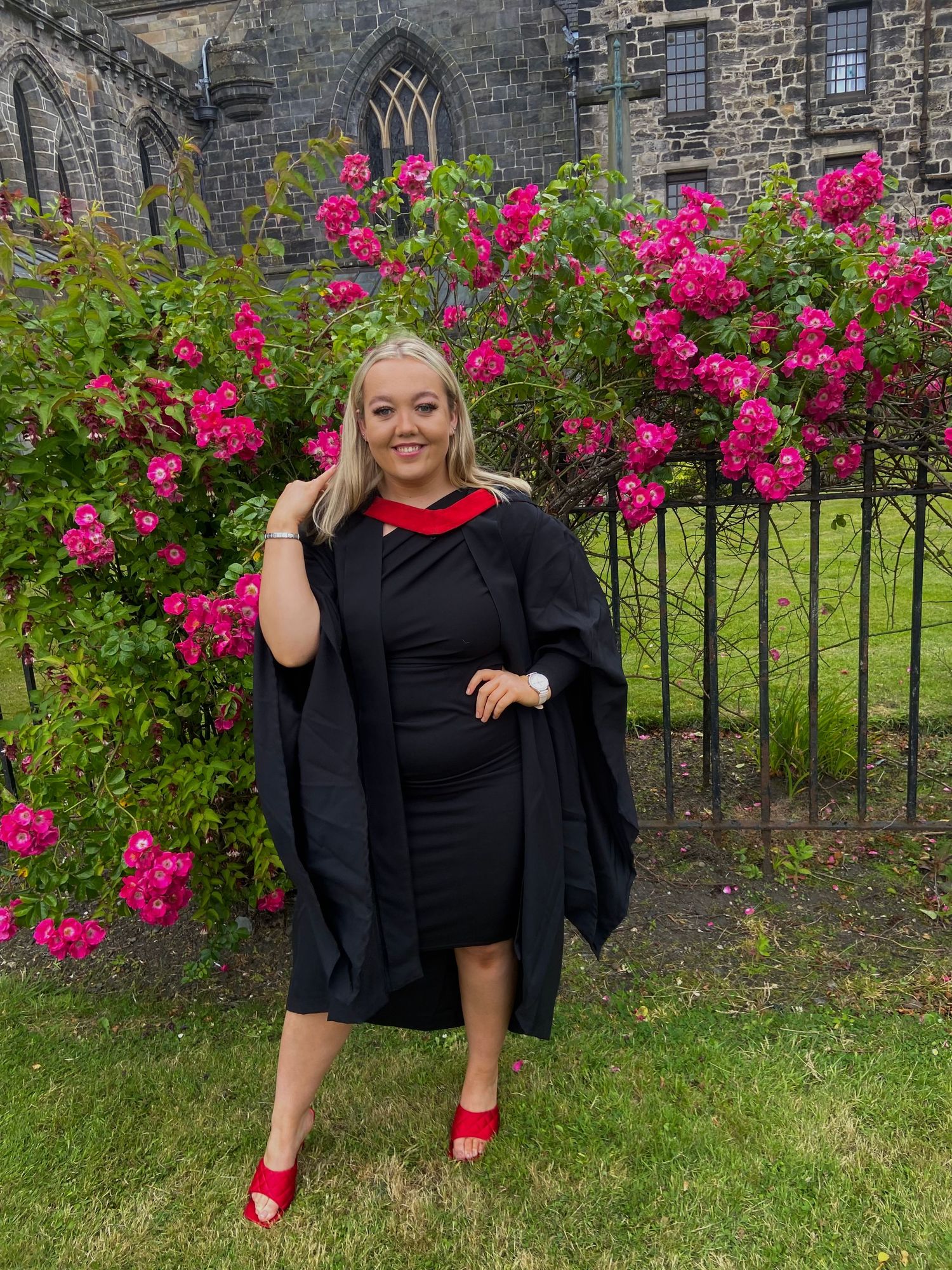 Graduate in academic gown and red heels posing in front of pink roses outside a stone university building.