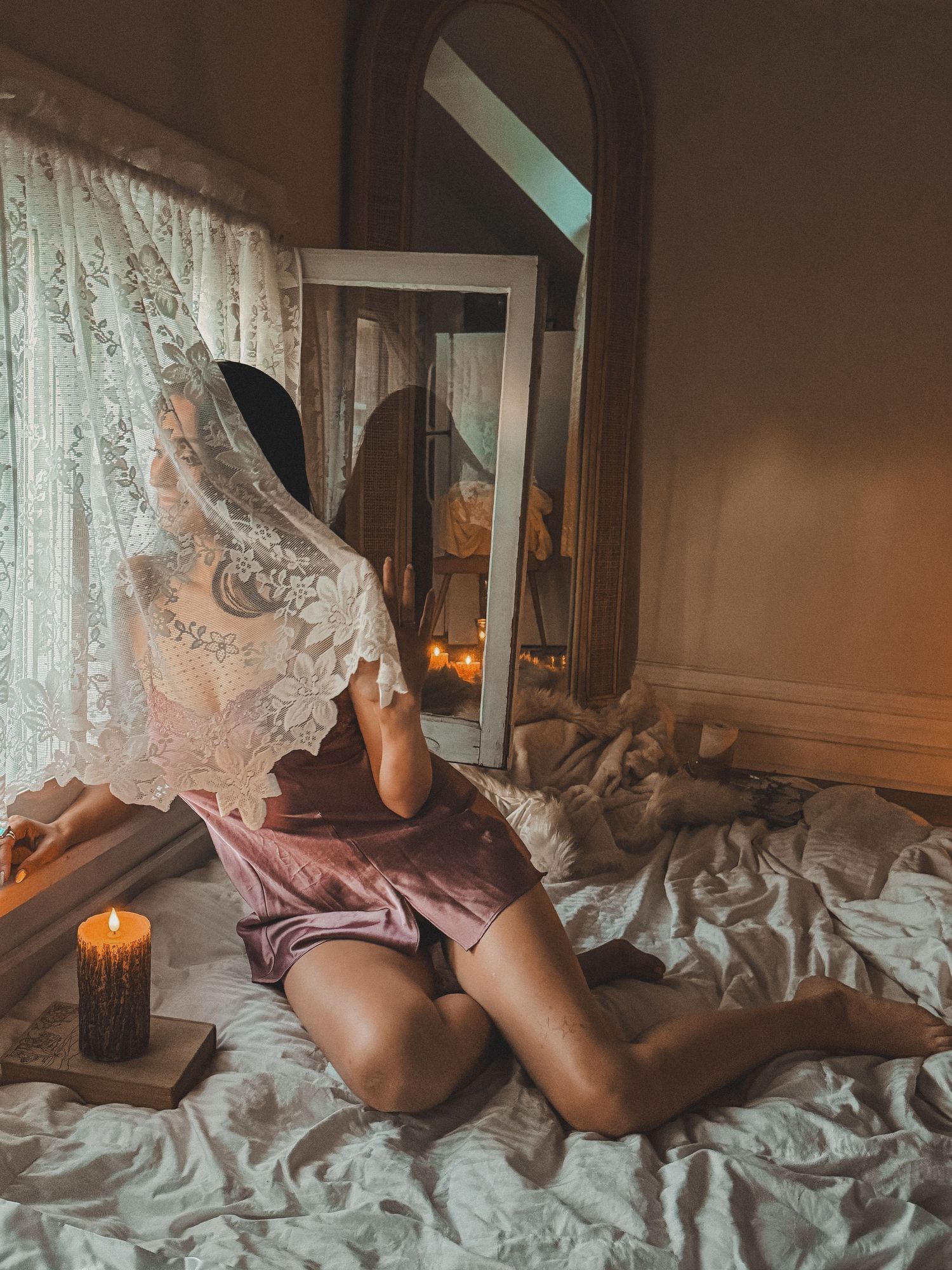 Woman in pink silk shorts posing on a bed with a lace veil, candle, and vintage mirror in warm light.