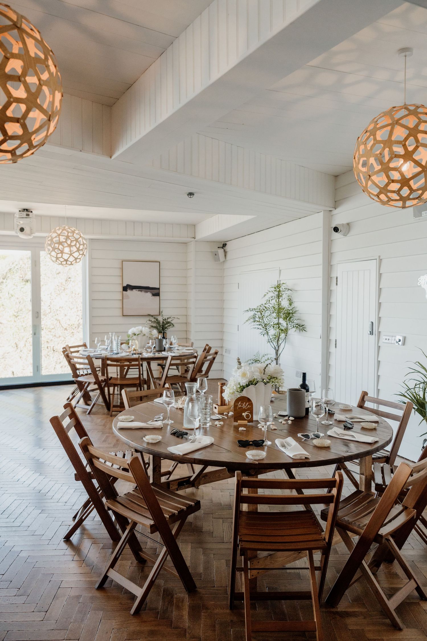 Bright dining room with round wooden tables, folding chairs, herringbone floors, and geometric woven pendant lights for an elegant event.