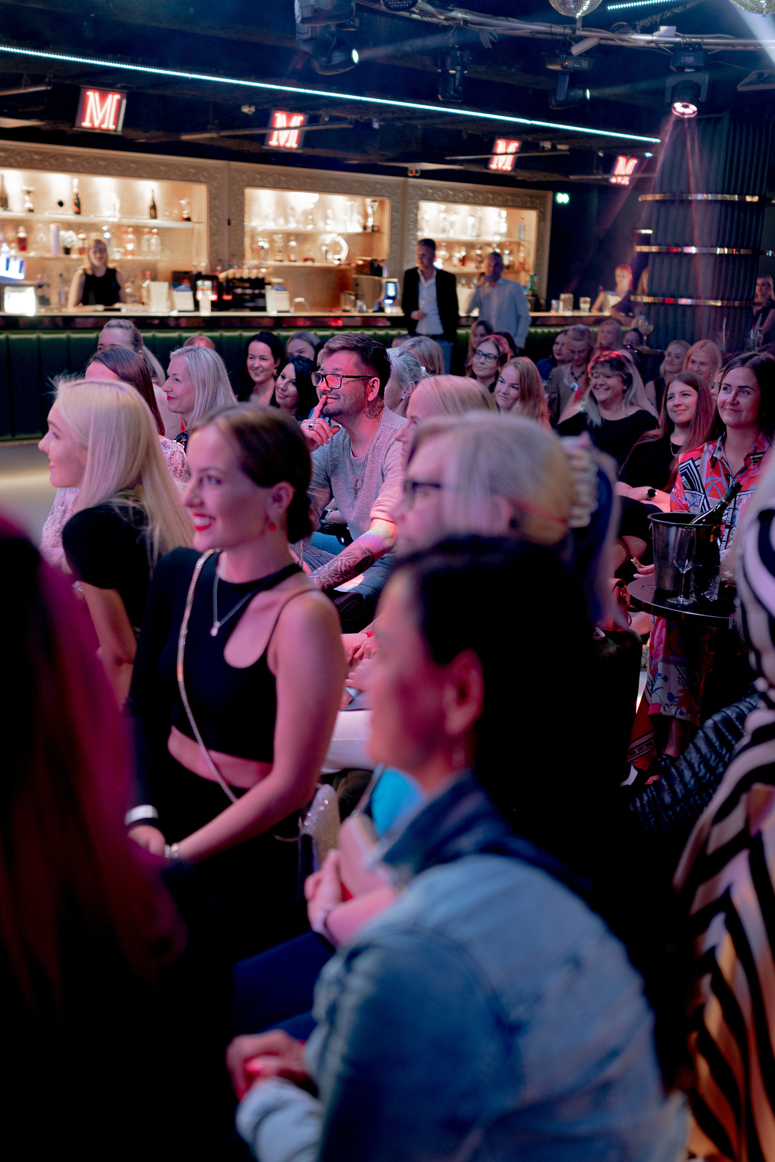 A crowd of people gathered at a trendy bar or venue for an event, with women in black outfits prominently in the foreground.