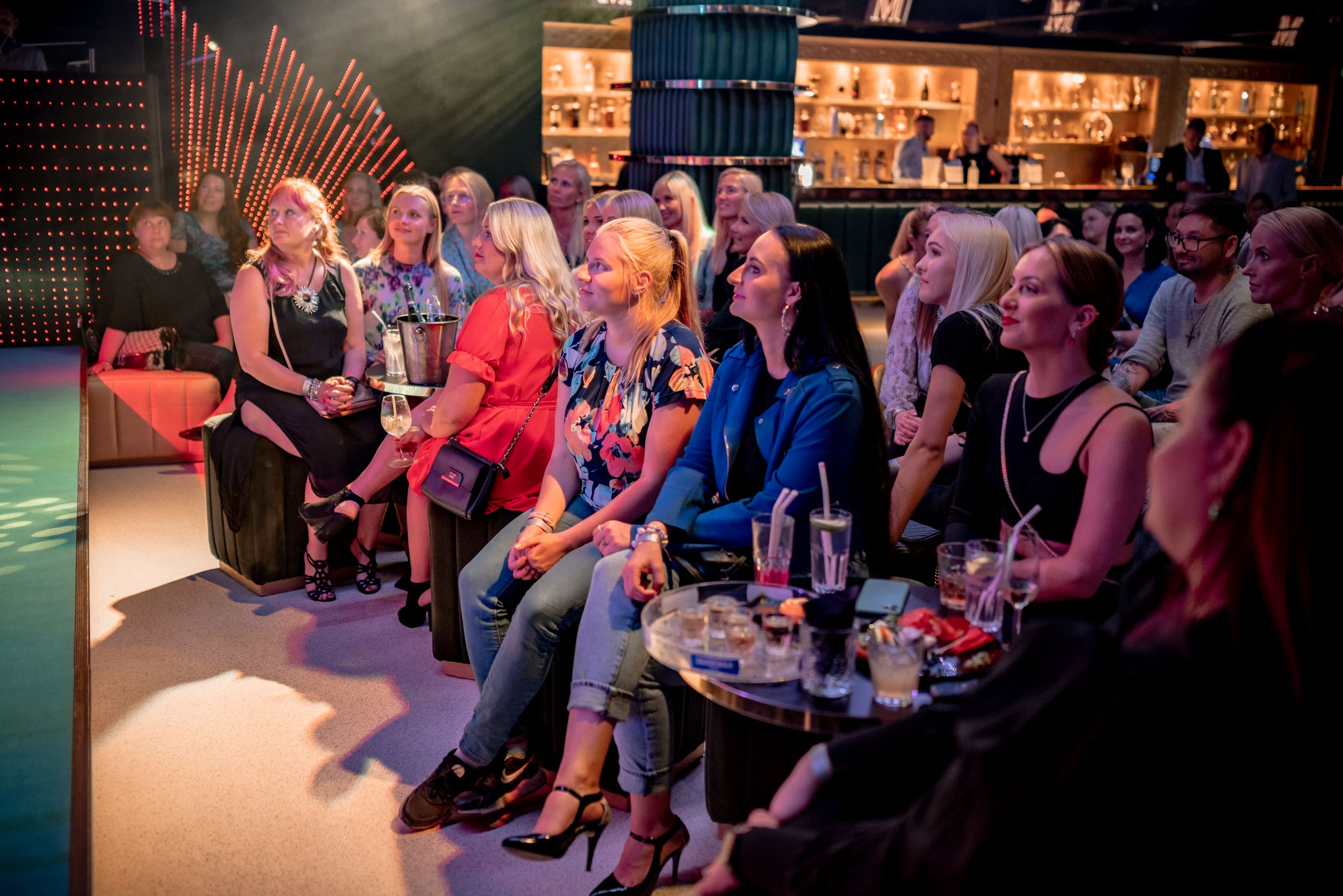 Diverse group of women seated at a bar venue, attentively watching an event presentation in a modern lounge setting.