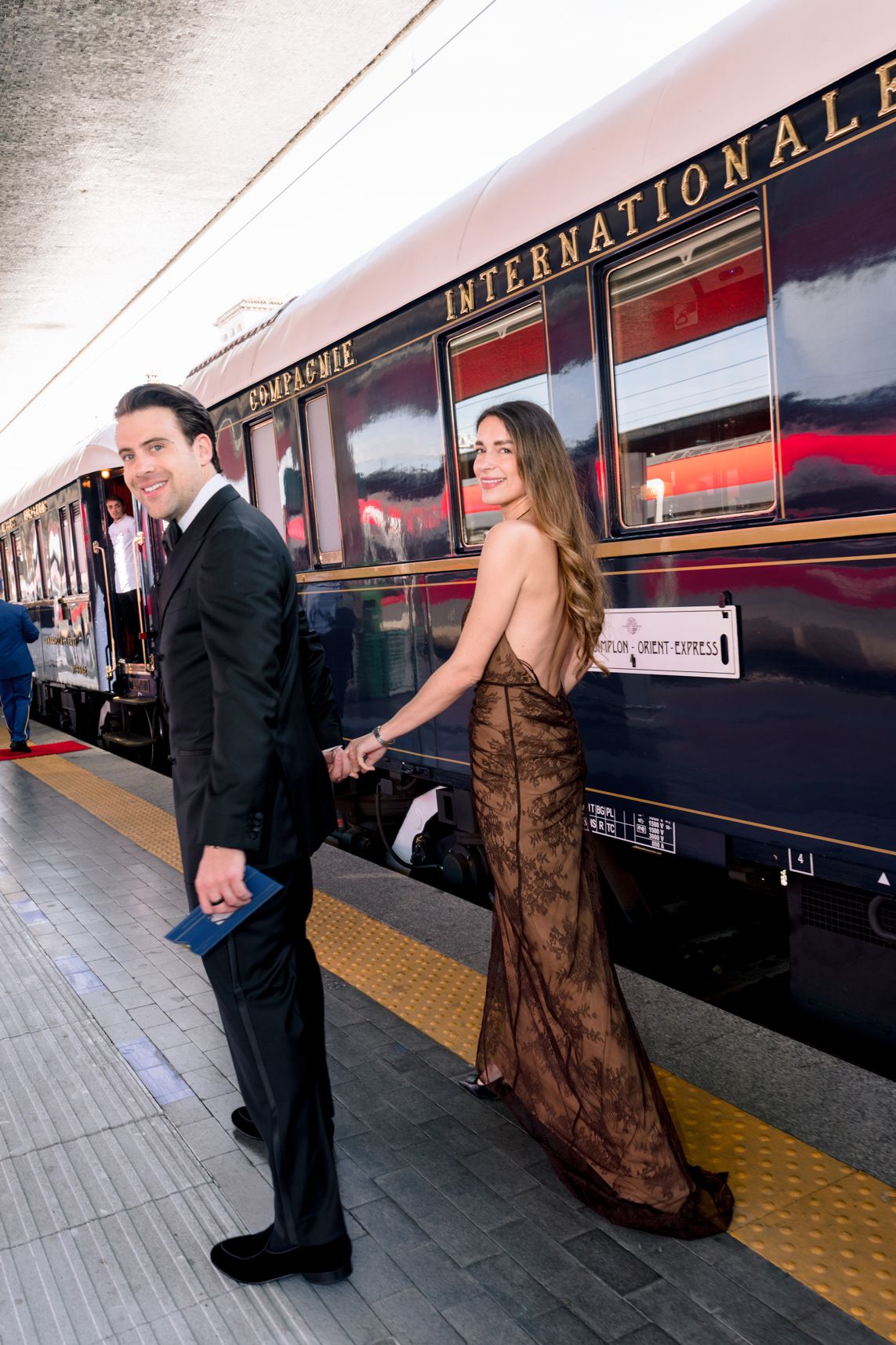 Elegant couple in formal attire holding hands on a train platform beside the Venice Simplon-Orient-Express.