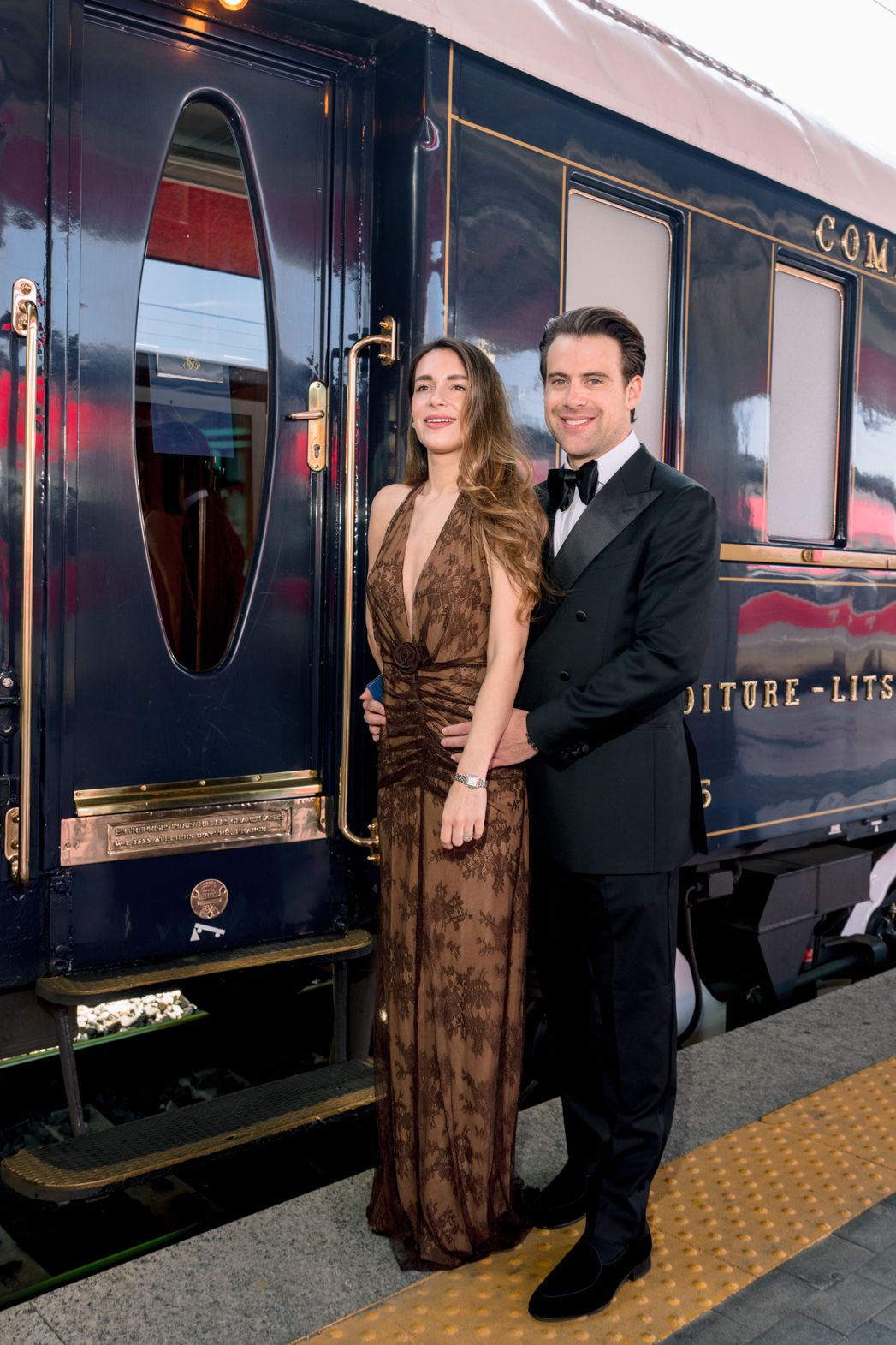 Elegant couple in formal attire posing beside a luxurious navy blue Orient Express train car on a platform.