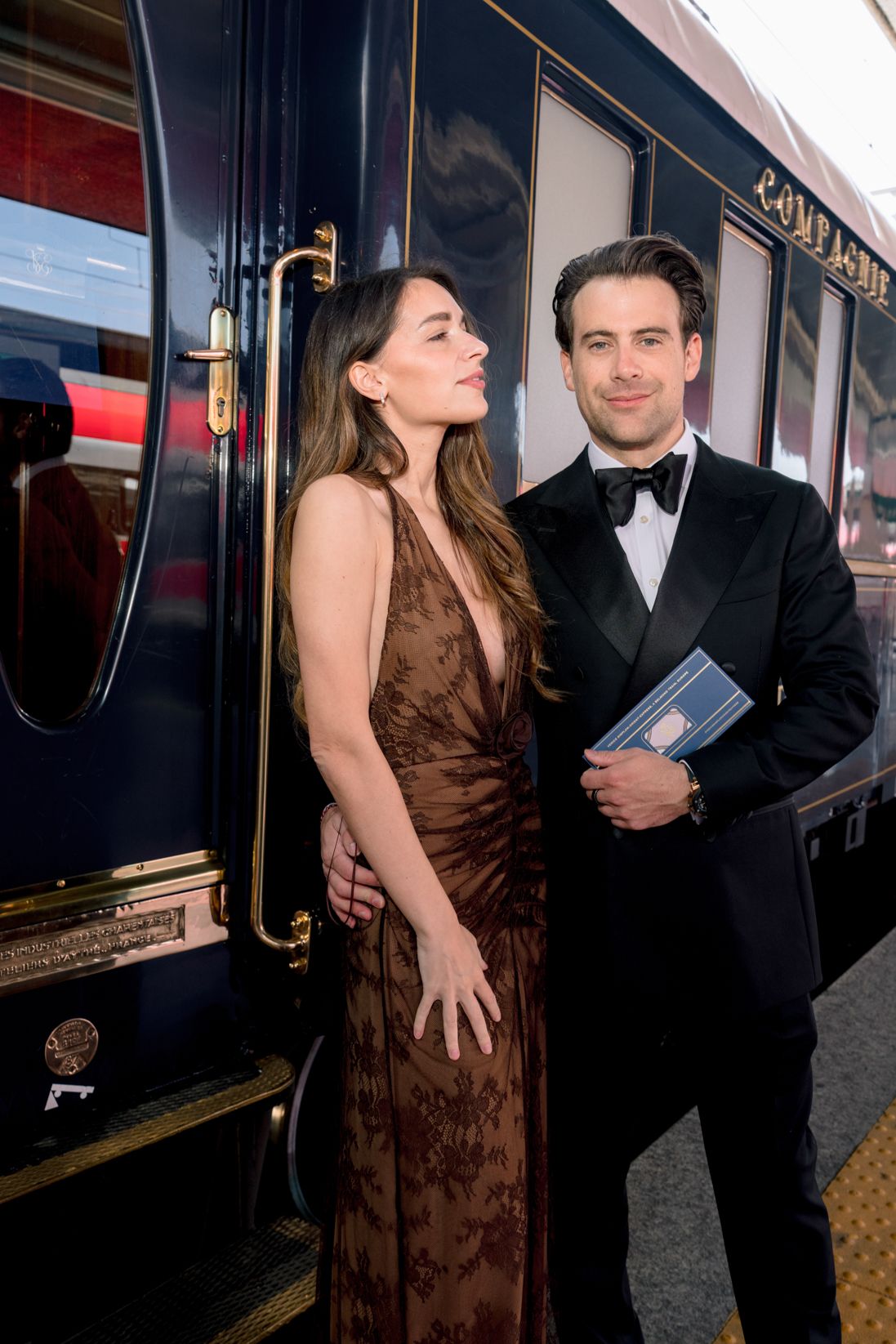 Elegant couple in formal attire posing beside a luxury navy blue train car at a station platform.