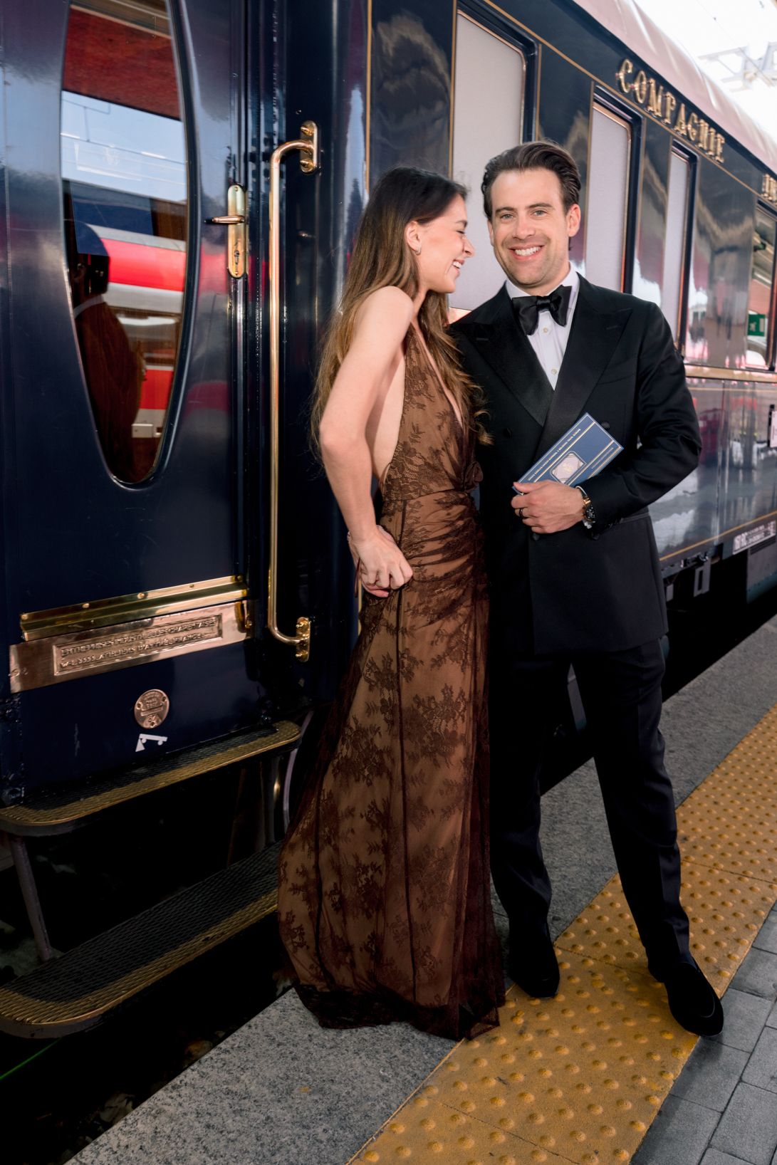 Elegant couple in formal attire smiling beside a luxury blue train car on a station platform.