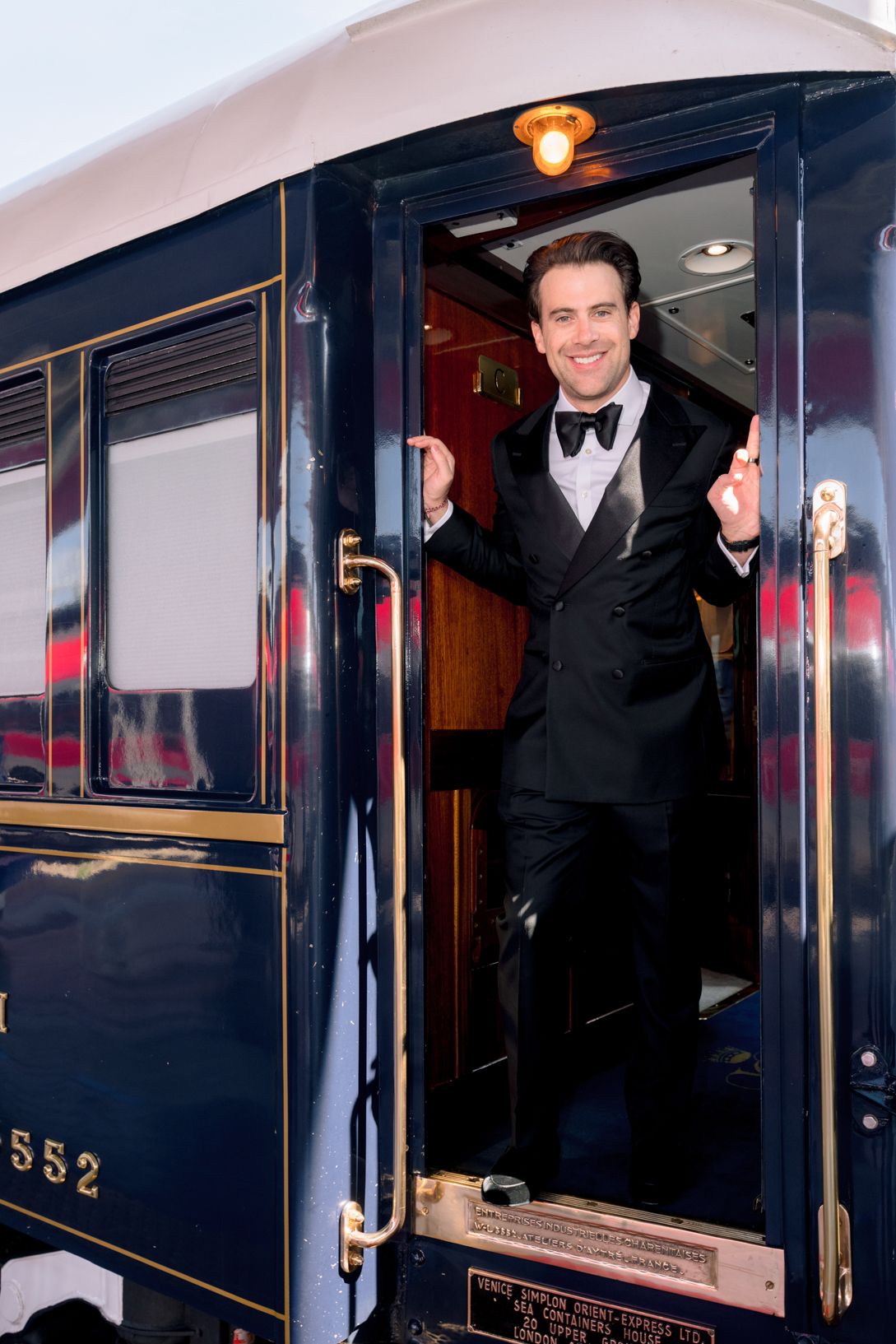 A smiling man in a black tuxedo stands at the open door of a vintage navy blue train car with gold trim.