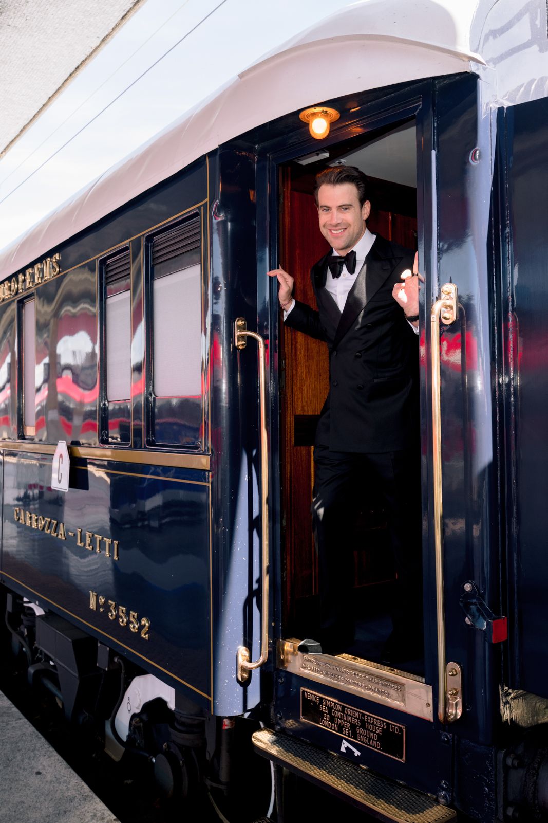 Smiling man in tuxedo standing at open door of luxury blue Orient Express train car number 3552.