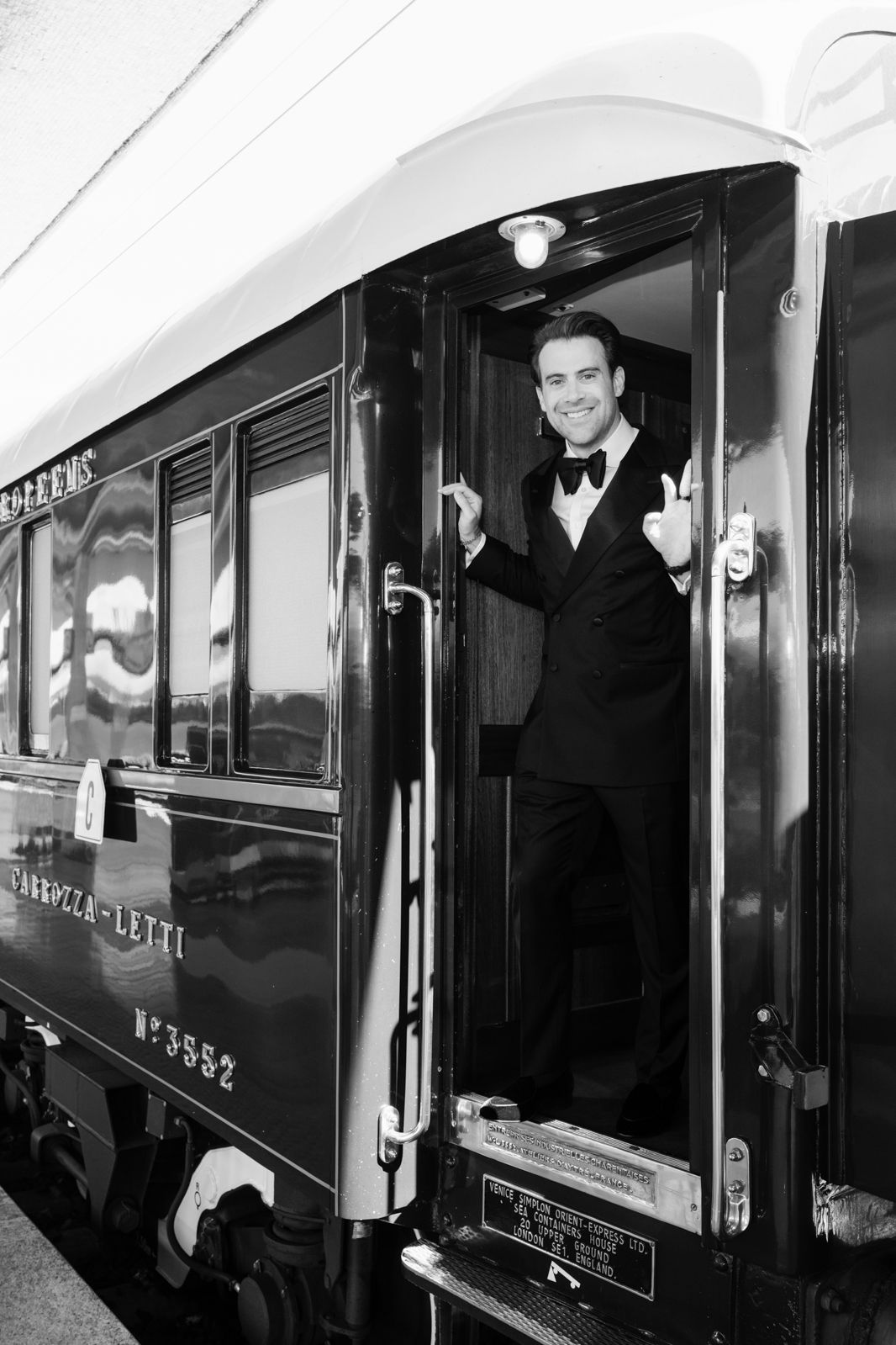 Smiling man in tuxedo standing at vintage train car door in black and white photo.