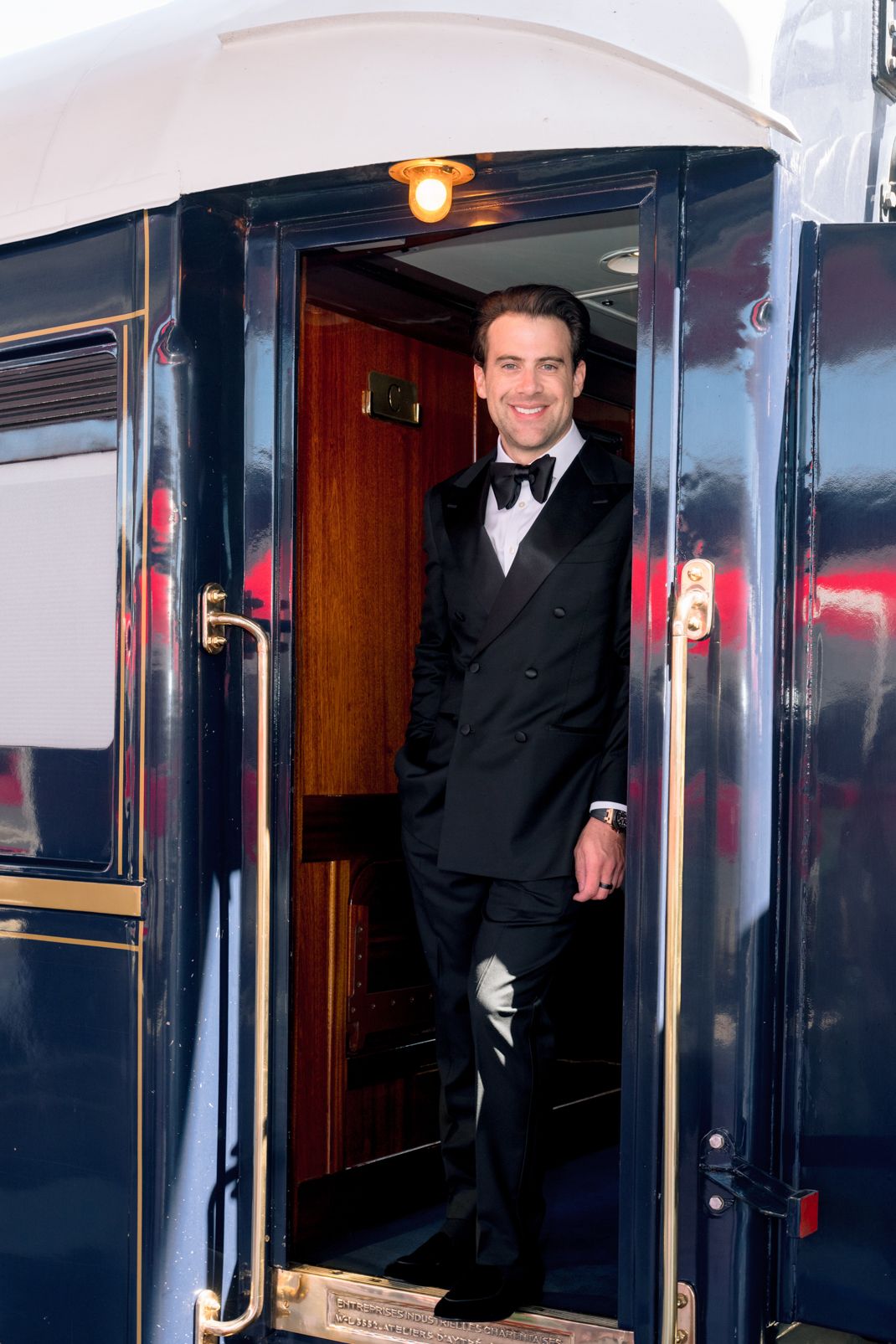 Smiling man in black tuxedo standing in doorway of luxury Orient Express train car.