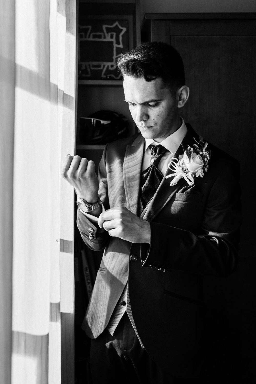 A groom in a formal suit adjusts his cuff while standing by a window in dramatic black and white lighting.