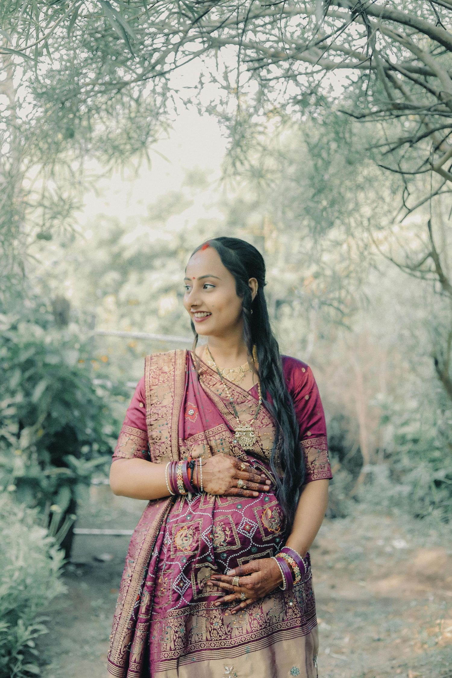 Pregnant Indian woman in pink silk saree with gold embroidery and bangles, smiling outdoors among green trees.