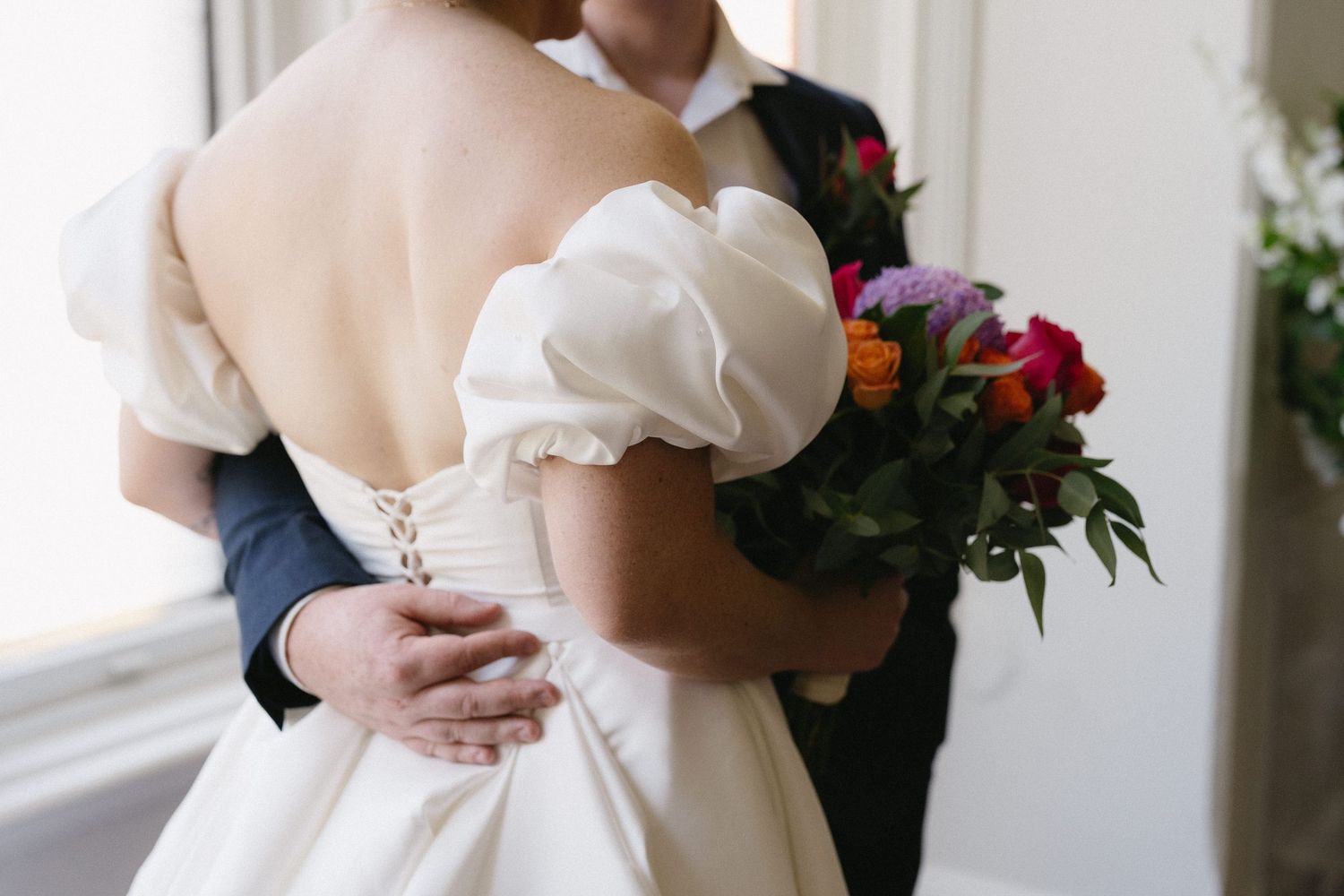 Bride in elegant off-shoulder white wedding gown holding colorful bouquet, embraced by groom in navy suit.