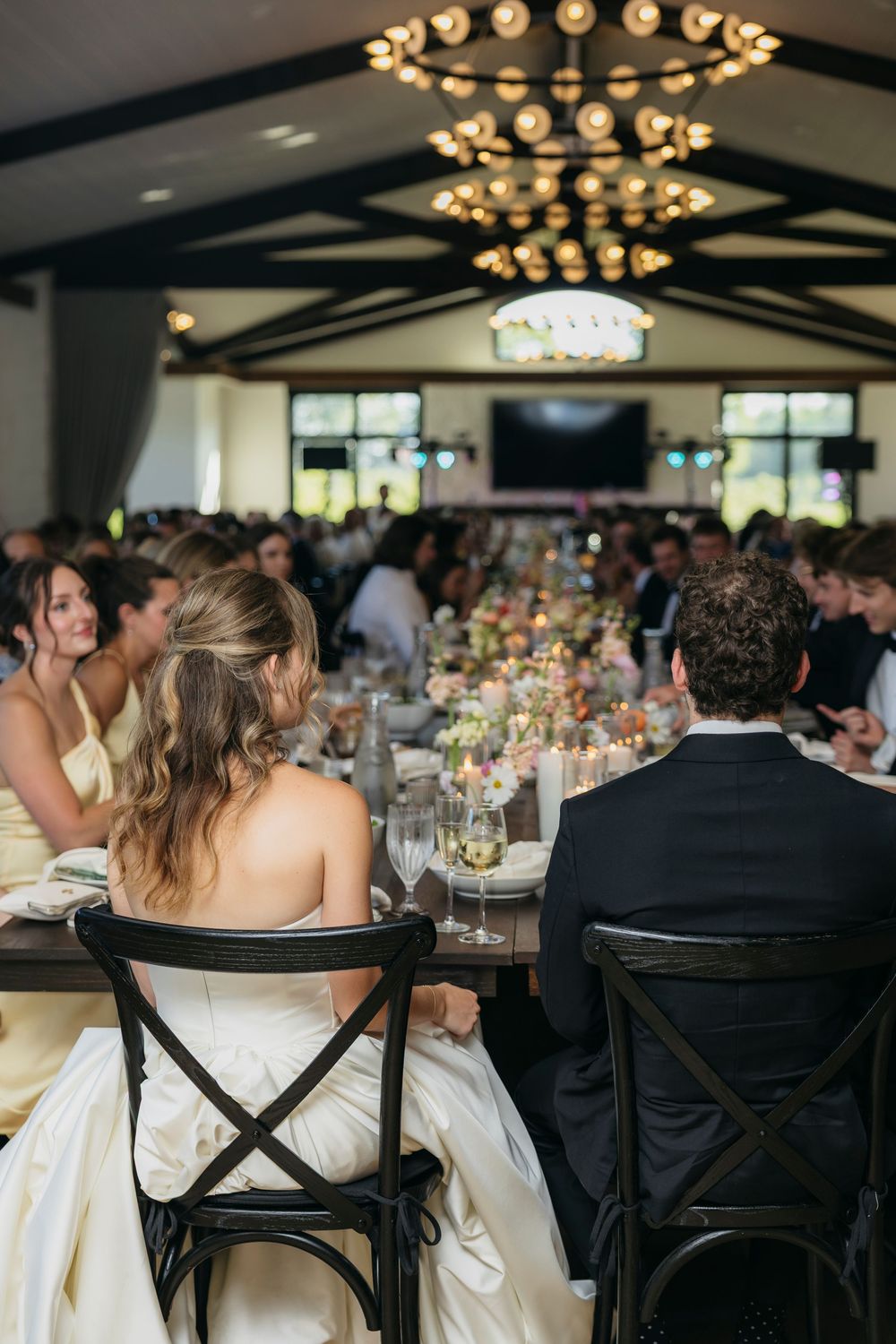 Bride and groom seated at elegant wedding reception with floral centerpieces and chandeliers captured by candid photographer in Wisconsin