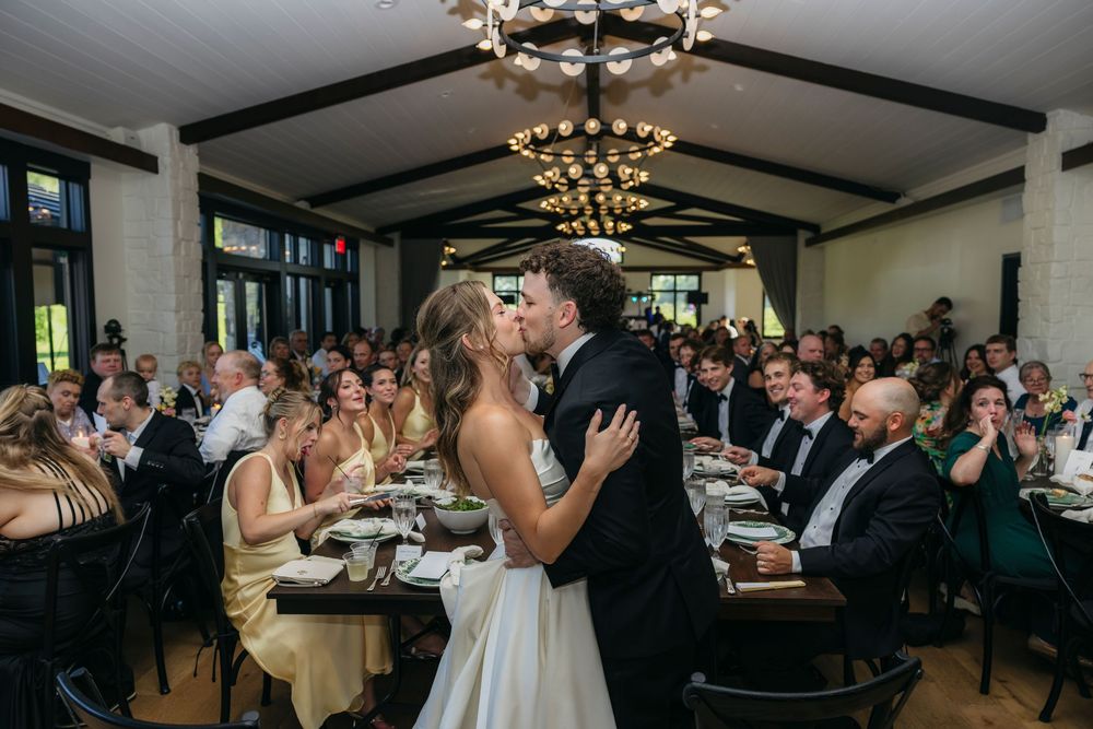 Bride and groom share kiss at elegant indoor wedding reception with guests seated at long dining tables captured by candid photographer