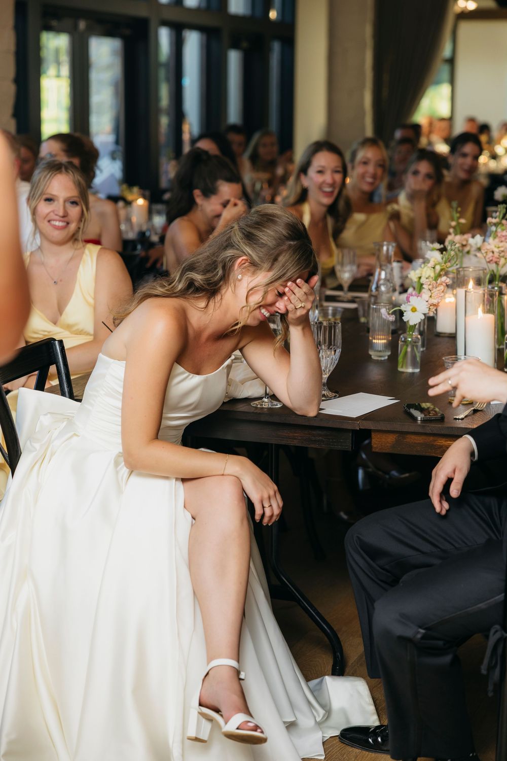 Bride in white gown laughing during wedding reception speech as guests smile in background captured by candid photographer in Wisconsin