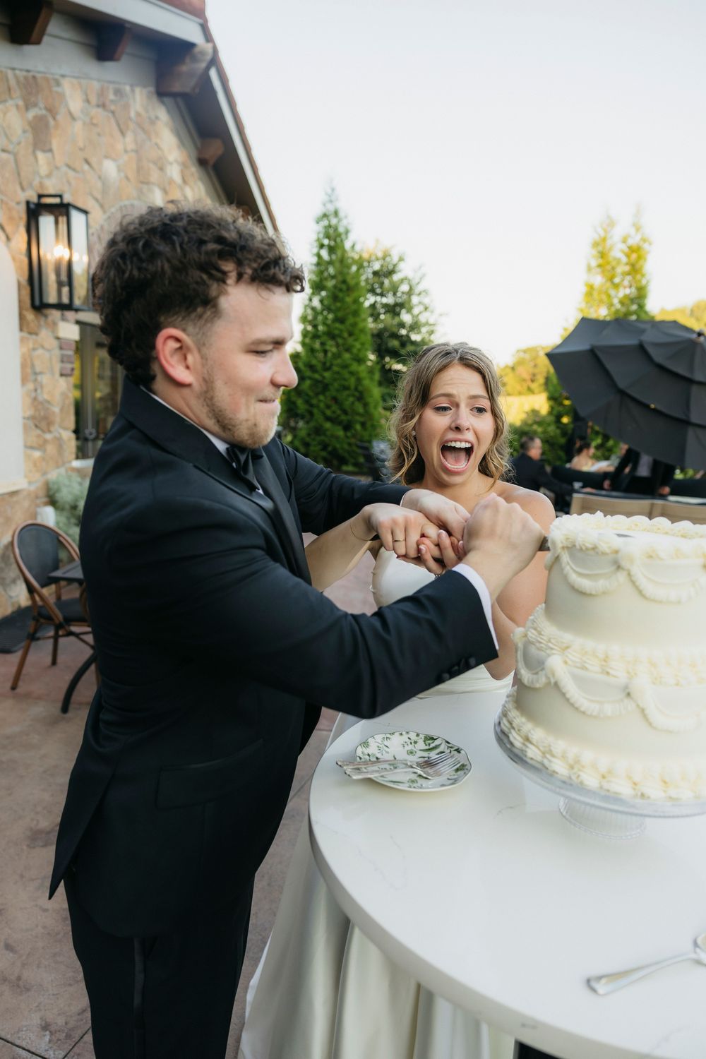 Bride and groom laughing while cutting their white wedding cake together outdoors at a stone venue reception captured by candid photographer