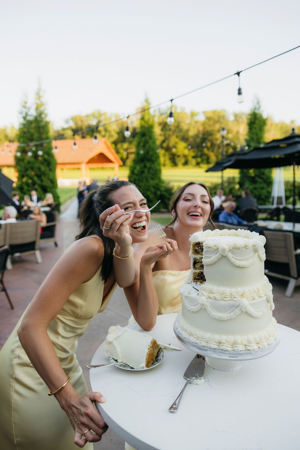 Two bridesmaids in yellow dresses laughing joyfully beside a white wedding cake at an outdoor reception captured by candid photographer