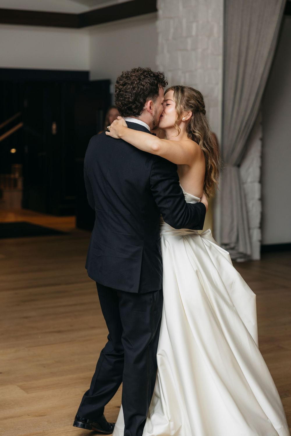 A bride and groom share a romantic first dance she in a white gown, he in black tuxedo, on captured by candid photographer in Wisconsin